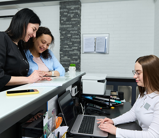 Two women at front desk with team member typing on computer